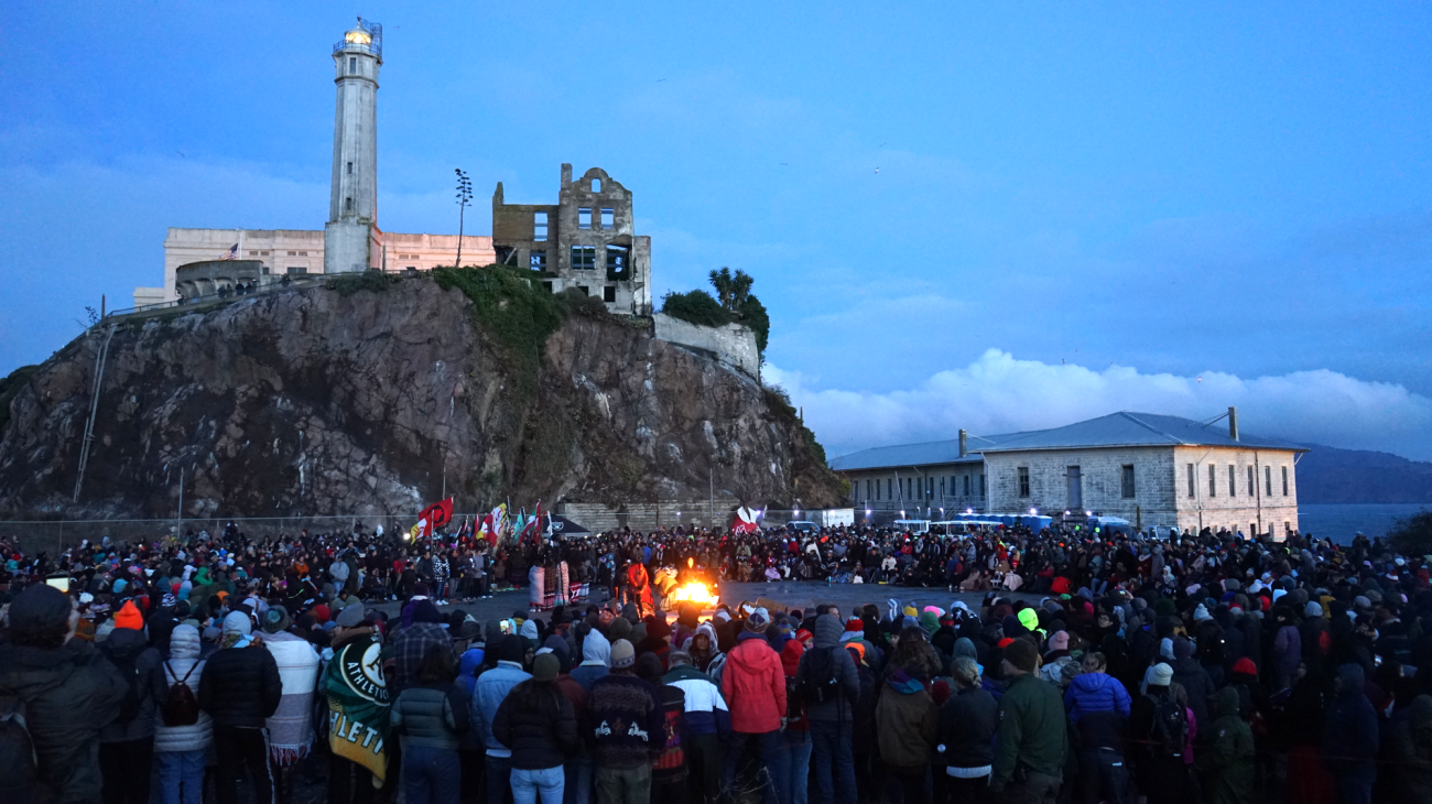 A big crowd of people surrounds a central bonfire, with parts of the Alcatraz penitentiary on a hill in the background.