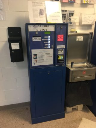 A clothing and e-cigarette dispenser next to a water fountain in a jail.