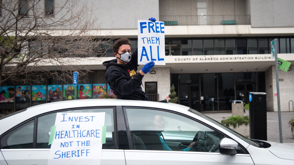 A protestor in sweatshirt, cloth sick mask, and dark gloves holds a sign out the sunroof of a car, reading "FREE THEM ALL."