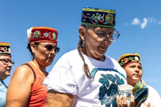 Four figures stand in front of a bright blue sky, heads bowed. 