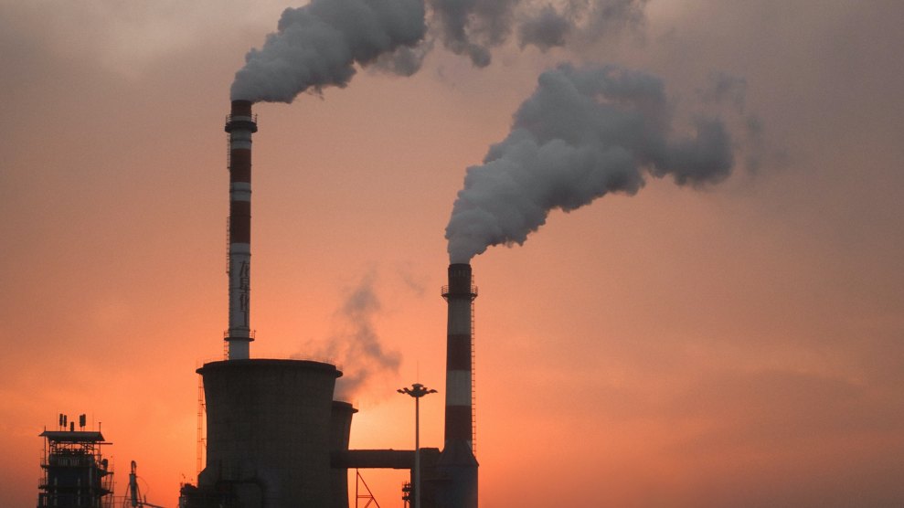 A photo of smoke coming into the sky around a factory. Due to a sunset or fire, the sky has an orange tint.