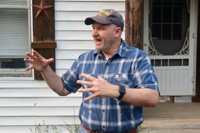 A white man in a blue checked shirt and a hat talking passionately in front of a white house