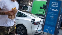 A man wearing shorts and a t-shirt eating a burrito walks past a white Tesla plugged in to a public charging station