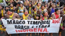 Indigenous people, in face paint and traditional clothes, marching with a banner that says, "Marco temporal"