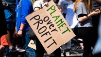 A white person holding a cardboard sign that says, "Planet Over Profit"