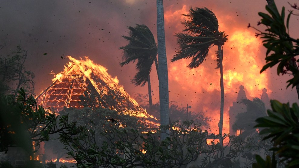 Orange and yellow wildfire engulfs a triangular building. Blackened trees lay in front.