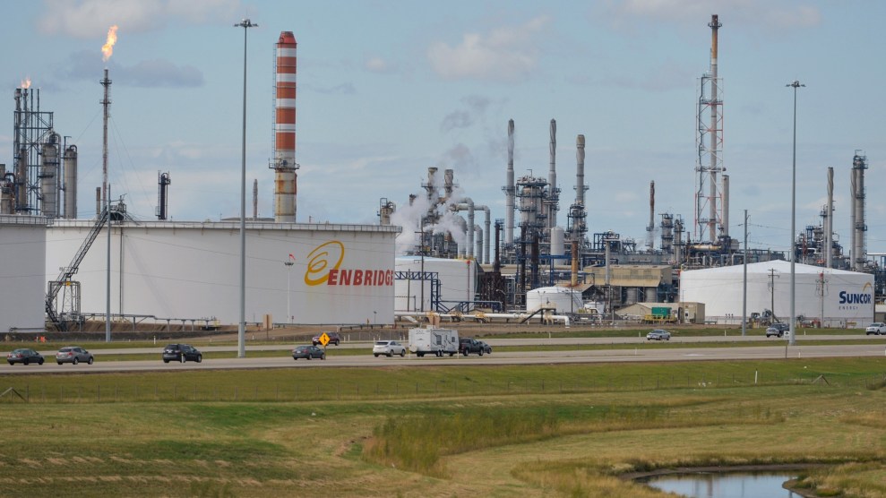 Behind a wetland, an oil refinery dominates the sky. It is made of mostly white metal and many smokey pillars.