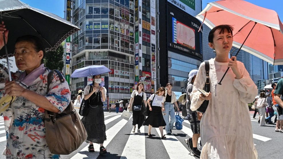 Several people stand in bright light at a sunny crosswalk. They use umbrellas and wear masks.