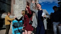 Two white youths raise their fists into the air in celebration. They stand next to a bright blue sign that partially reads "Montana Youth Making." In the background, others give high fives.