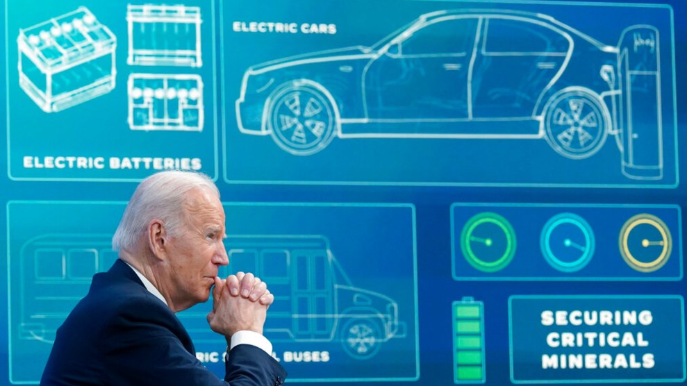 Joe Biden sits with his chin in clasped hands in front of a blue board projecting information about electric vehicles, including an outline of a car and battery and the words "Securing Critical Minerals."