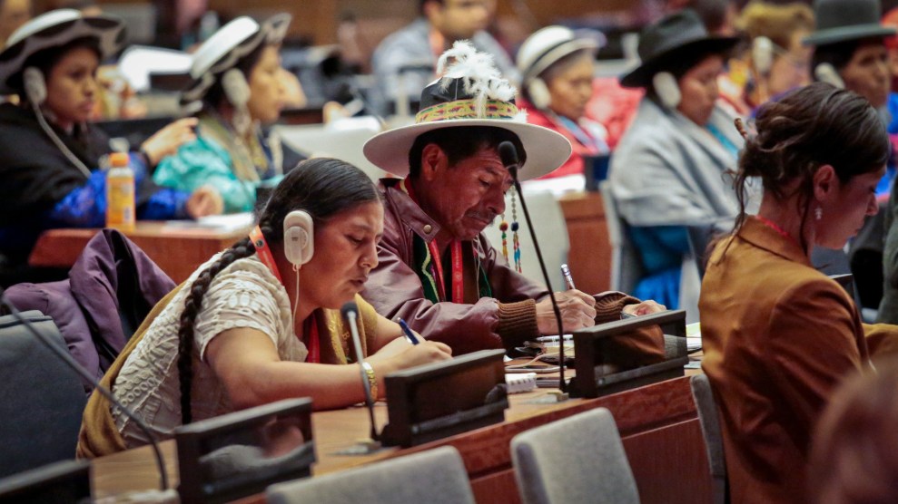 People sit at tables facing one direction. The camera is focused on two individuals with dark hair. One is braided and one is cropped short. Many people wear hats with brims.