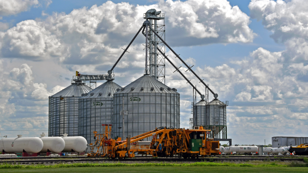 Grey grain storage silos tower along side the railroad tracks in rural Lebo, with white storage tanks of liquid ammonium nitrate fertilizer crowding the bottom. The sky is light blue with long fluffy clouds.