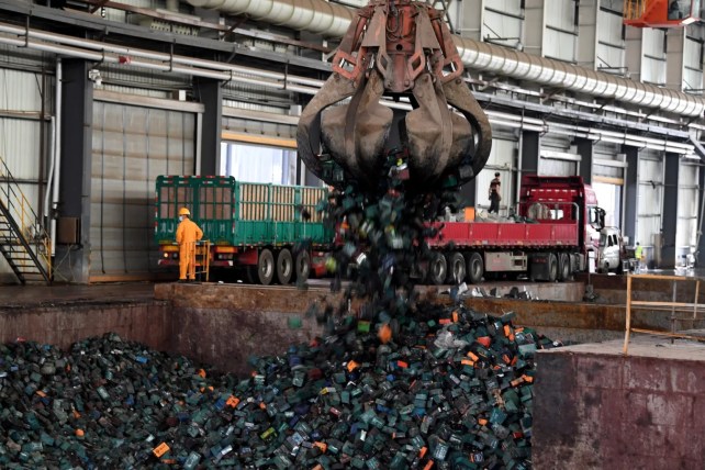 A large claw machine drops black batteries into a pile. A worker in a bright orange suit stands behind the pile.