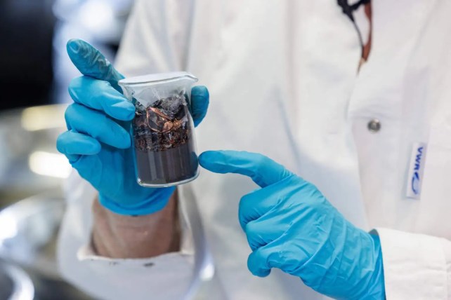 Blue-gloved hands hold a glass beaker filled with brown and black material.