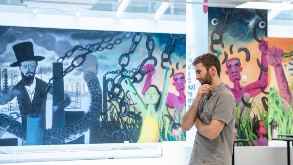 A white person with brown hair and a beard stands in front of an art exhibit. Behind them are large brightly colored murals.