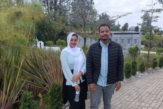 A woman with light brown skin in a hijab, smiling, and a man with darker brown skin and black hair also smiling, with some bushes behind them.