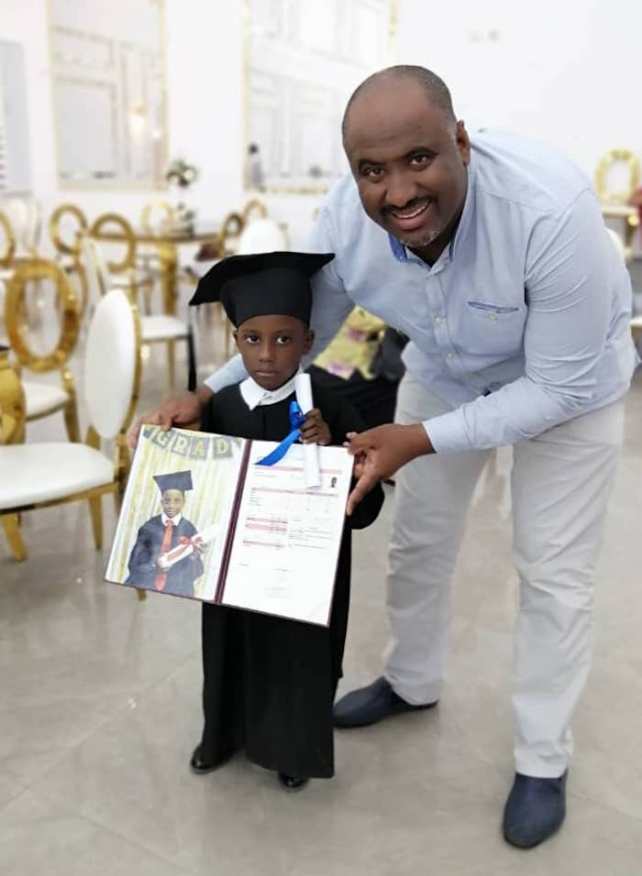 A man with dark brown skin with his son, who is a few years old, in a cap and gown, holding a certificate and photo of the son