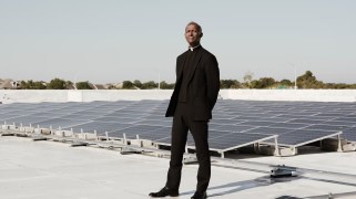 Pastor Antoine Barriere, a Black man dressed in all black, next to solar panels