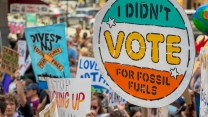 A crowd of protesters hold signs, including one, in focus, that says "I didn't vote for fossil fuels."