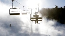 A ski area featuring a ski lift with someone in it, someone skiing, and what appears to be a lot of snow.