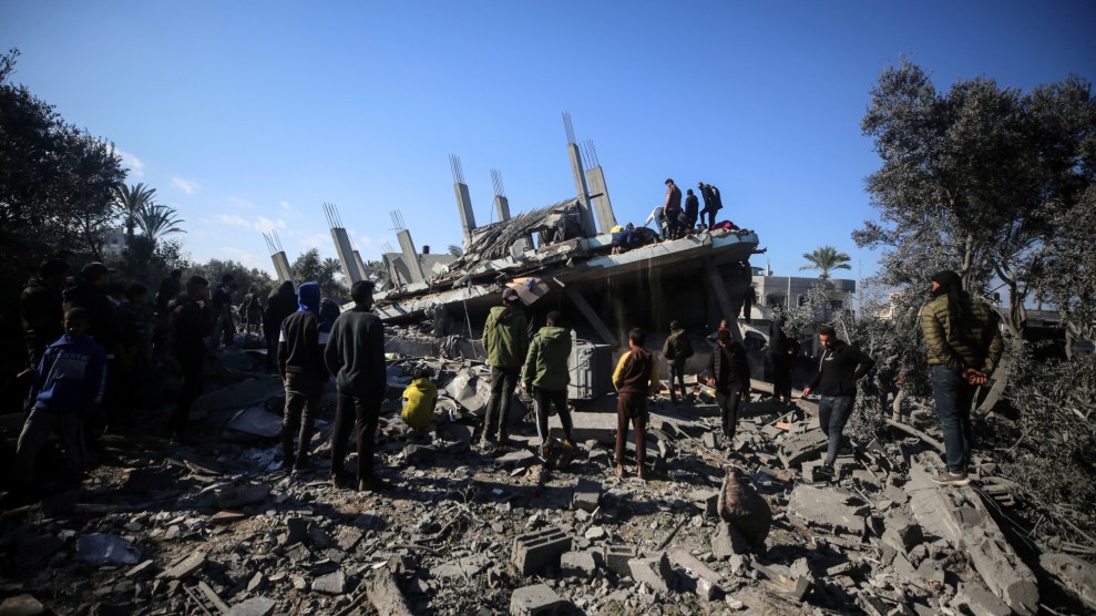 A group of people gathers in front of a pile of rubble. Trees covered in grey dust frame the photo.