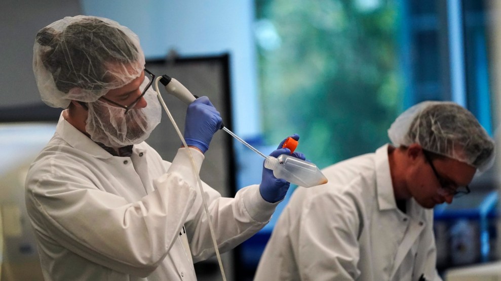 Two white individuals wearing white lab coats and hair nets look at their work in a blurred blue and green lab.