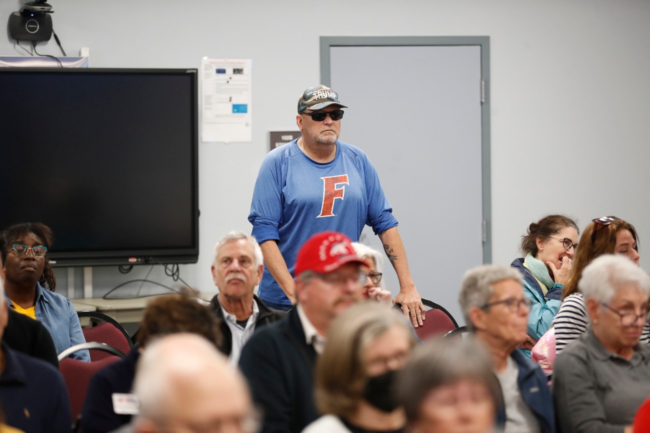 Man in sunglasses stands at the back of a room full of people.