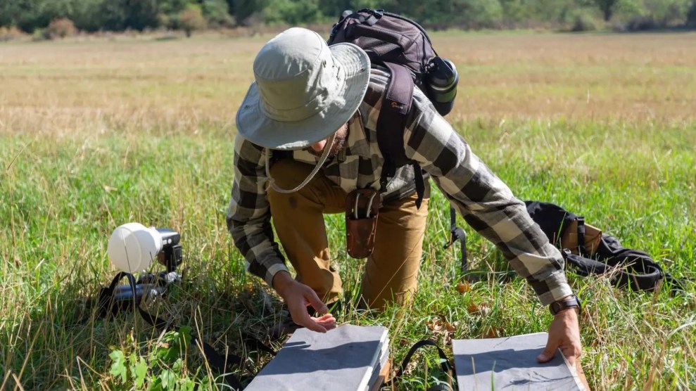 A white researcher with a brown beard and a tan hat covering their face kneels on a grassy field.