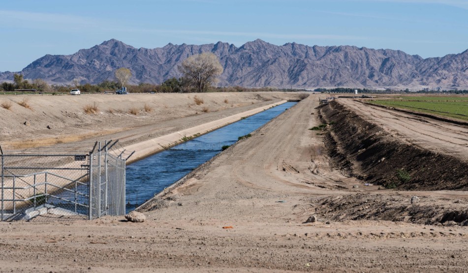 A blue strip of water cuts through a light brown desert. In the distance mountains loom.