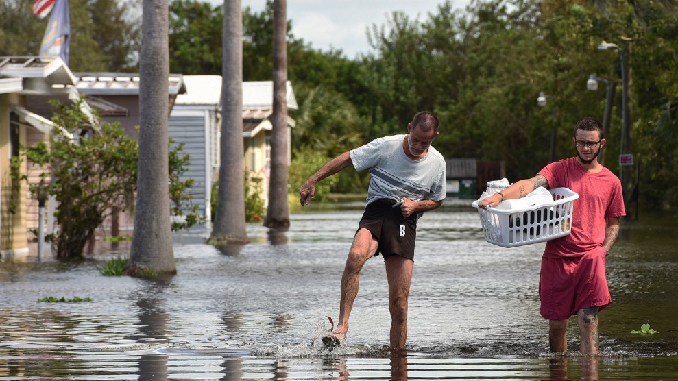 Two men walking in flooded streets with some houses behind them, with one carrying a laundry basket