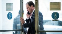 A white man walks through a door, speaking on the phone. He wears a suit.