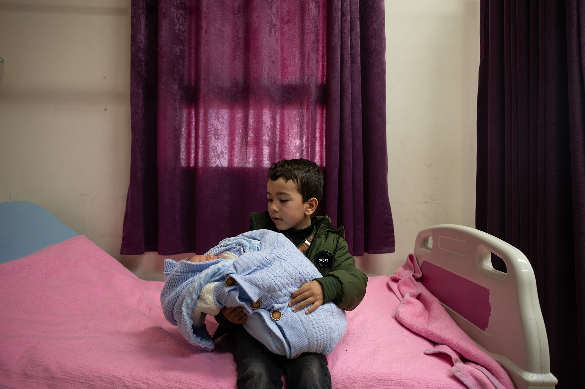 Young boy sitting on a hospital bed, holding a newborn.