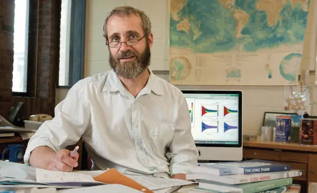 Portrait of bearded man sitting at a desk with papers on it.