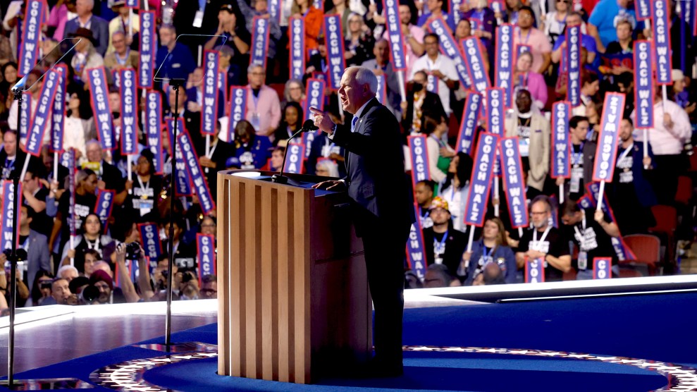 Tim Walz stands on stage behind the podium before a sea of blue 'Coach Walz' signs.