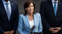 New York Gov. Kathy Hochul, a white woman with brown hair, wearing a light blue suit. She is speaking into a microphone and has two men, one white and one black, behind her in suits.