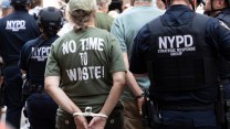 Outside the Tribeca home of Citibank CEO Jane Fraser, NYPD officers escorts a zip-tied protester wearing a green tee shirt that reads, on her back, "No time to waste!"