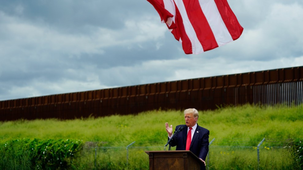 Donald Trump speaks at an outdoor podium, wearing a dark blazer and a red tie, against a section of unfinished border wall on the southern border, under an American flag.