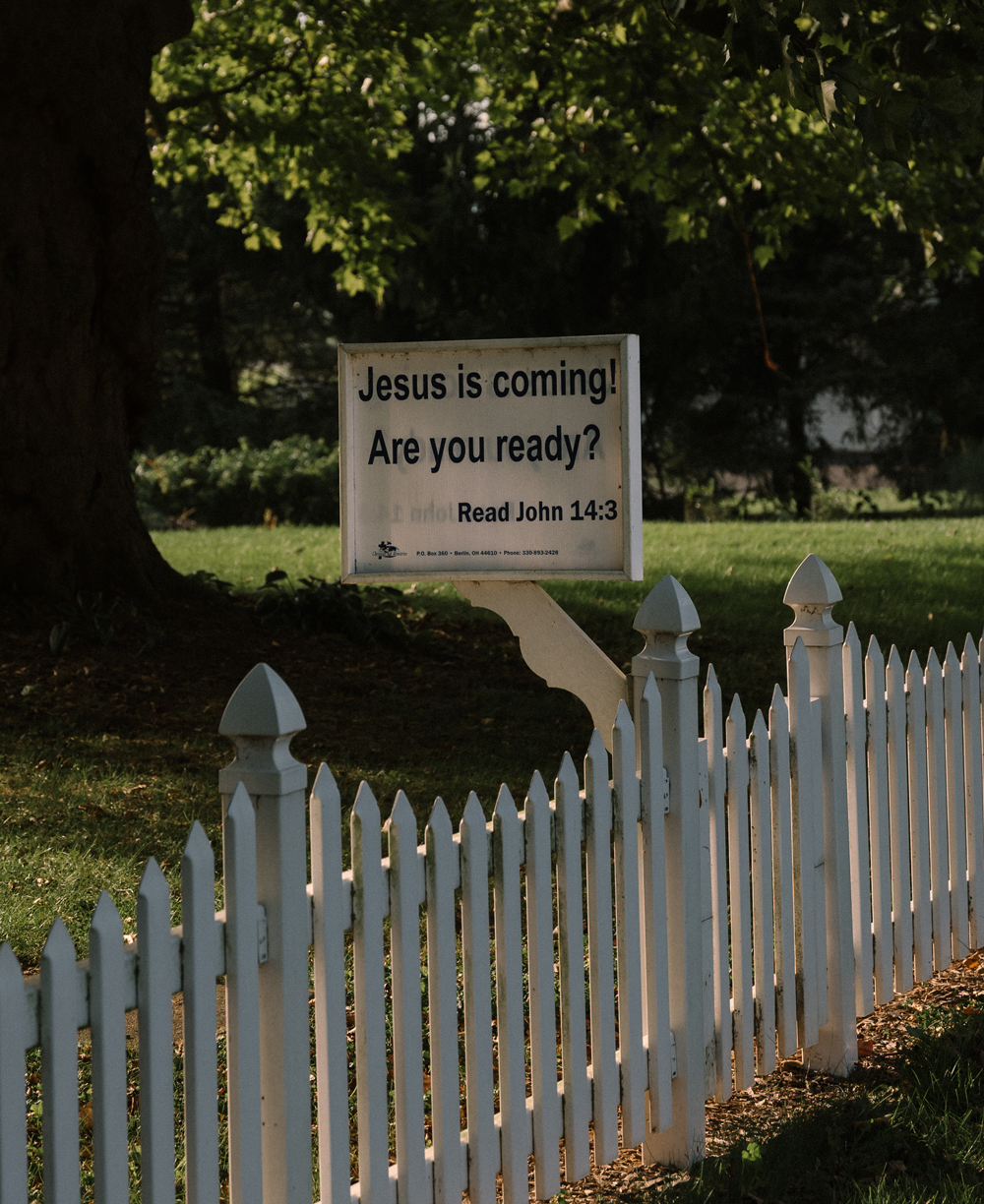 A white picket fence surrounds a yard where a sign reads, “Jesus is coming! Are you ready? Read John 14:3.”