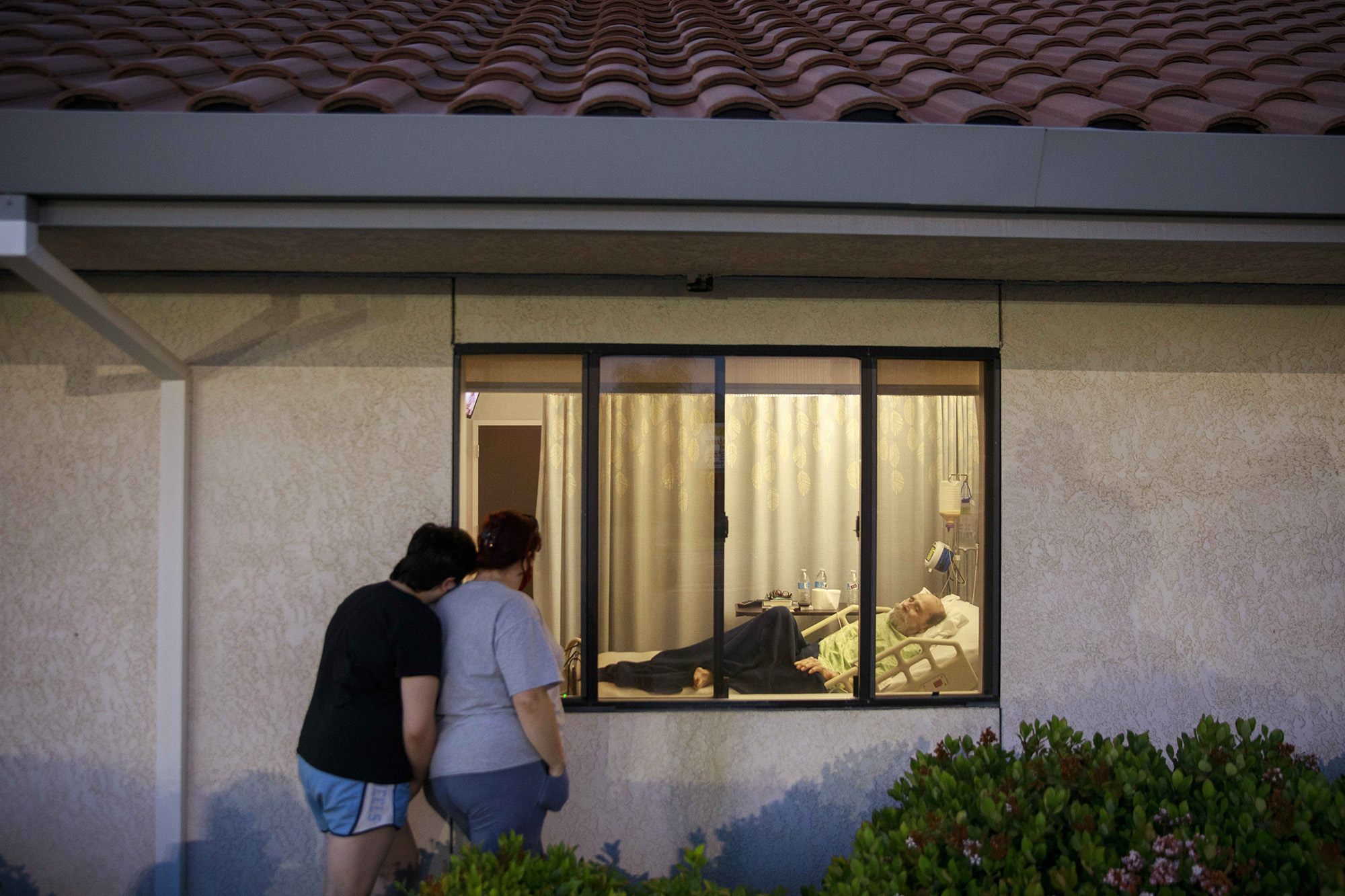 Long shot view of two people standing outside a medical facility, looking in the window at a man on a hospital bed.