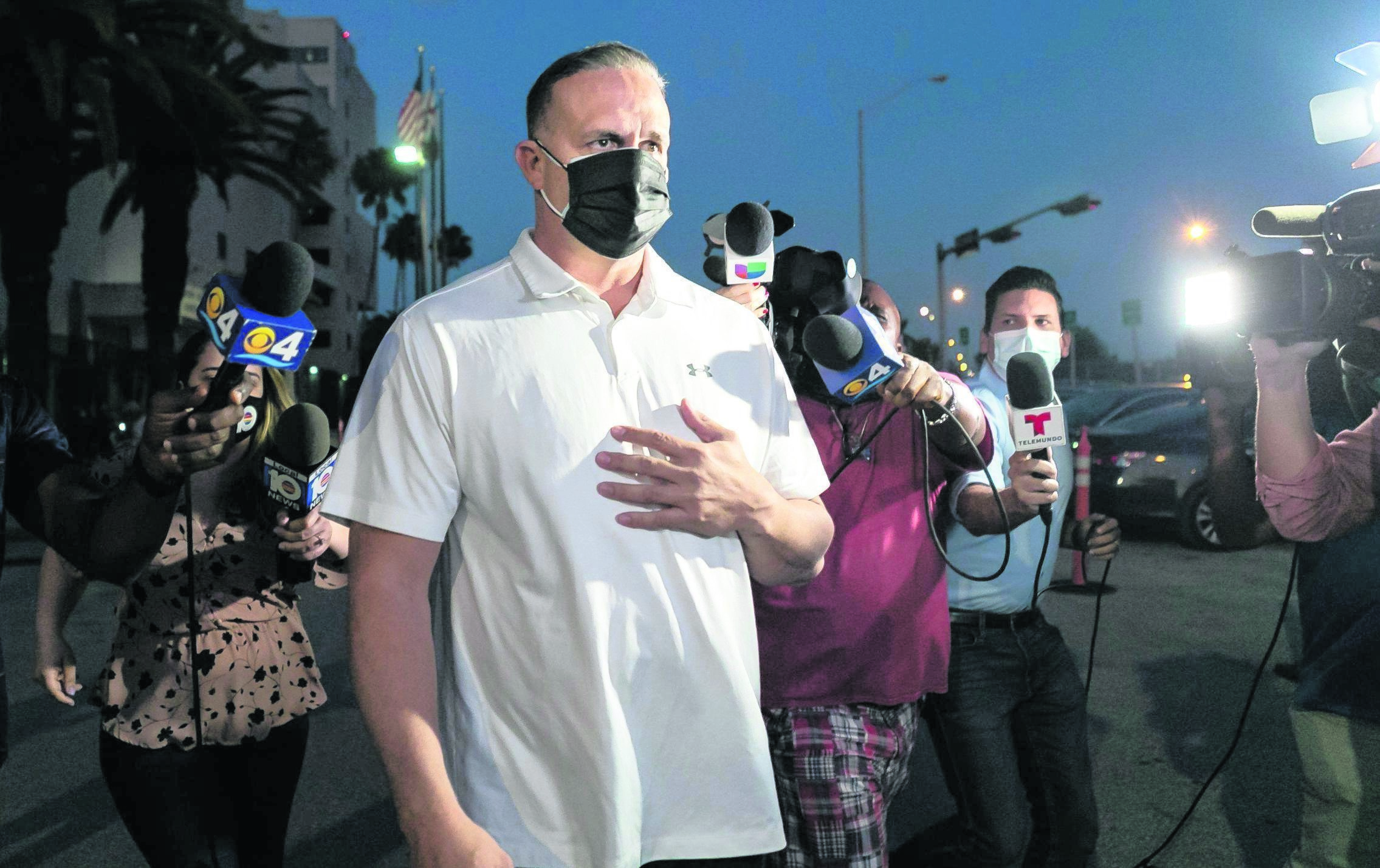 Man wearing a mask wearing a white shirt surrounded by TV cameras.