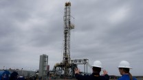 Two people in hard hats survey a drilling rig by Chevron under a grey, cloudy sky,