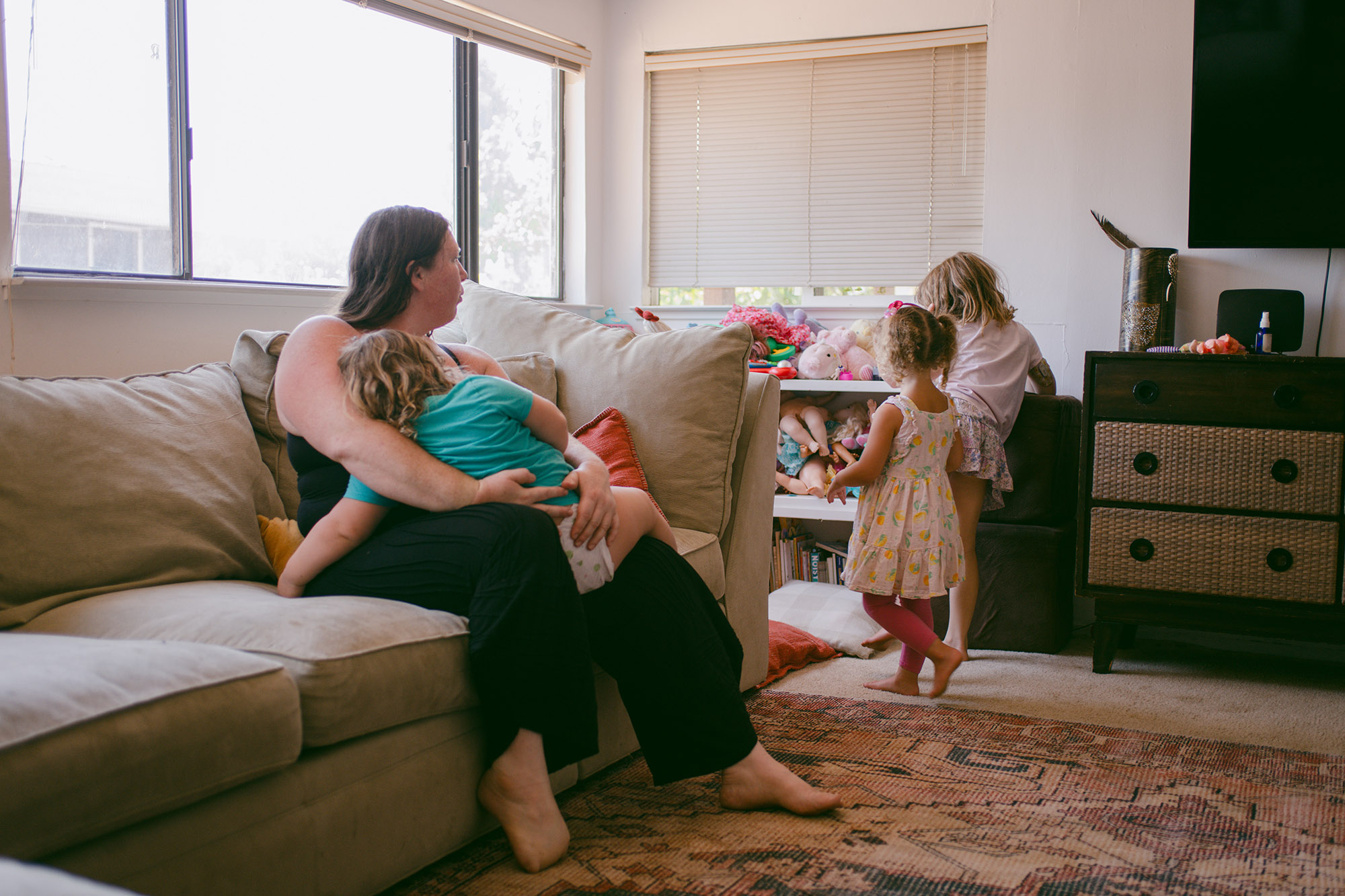 Susan Horton, a White woman wearing a black tank top and black pants, sits on a couch while she holds onto her toddler. She watches her other two daughters as they walk to their toys.