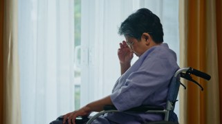 An aging person with light brown skin and dark gray hair in a wheelchair before the window. They have one hand over their face and are looking down. They are wearing a blue shirt.