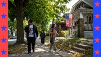 Two canvassers walk down a sidewalk. An American flag hangs from the porch of a home beside them.