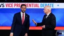 JD Vance and Tim Walz stand together on stage. Vance leans awkwardly in Walz's direction, as Walz holds up his right hand as if to steady Vance if needed.