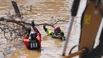 Two people in red and orange jackets, helmets and life vests are chest deep in muddy water, manipulating a fallen tree branch