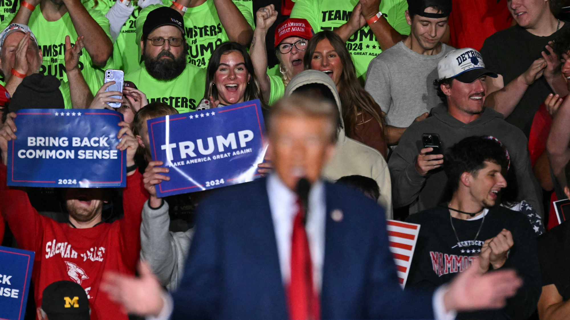 A photo of out-of-focus Donald Trump standing behind the podium, speaking to the crowd at a rally. Behind him, in focus, are people with pro-Trump signs, wildly cheering.