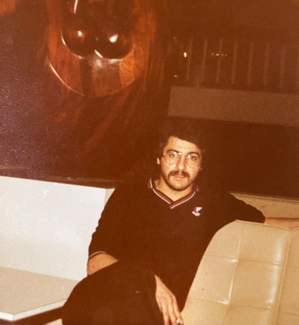 Ryan Khojasteh’s uncle Cyrus Salehi leans back on a sofa, with his left arm extended along the top. He sits with his right leg crossed over his left. His right hand in his lap, holding a cigarette.