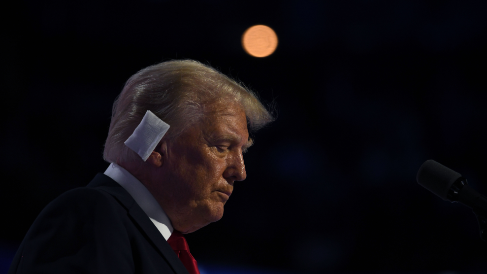 Donald Trump, seen in profile from his right in front of a bank of microphones, tilts his head downward with eyes closed. He has a bandage on his right ear.