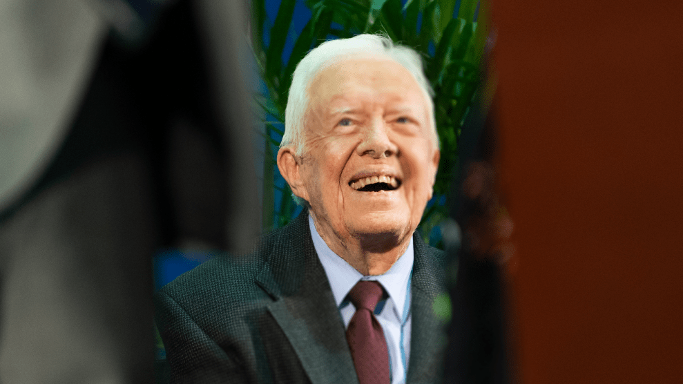 A picture of Jimmy Carter, an aging white man, wearing a suit. His face in this photo is framed between the bodies of two people in front of him. He is looking upward, with a smile on his face.